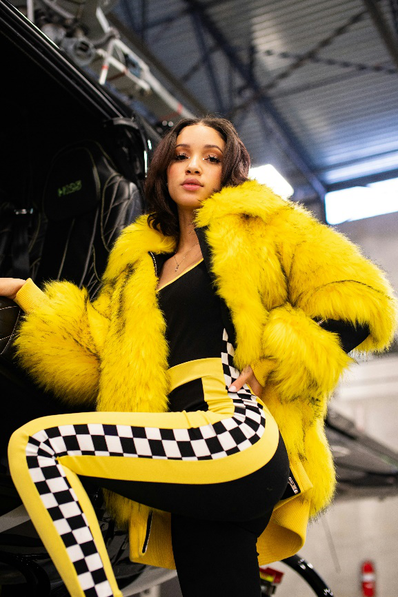 Woman posing by a vehicle in a vibrant yellow fur jacket.