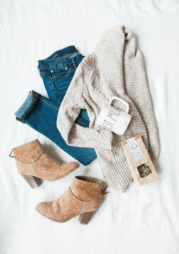 Flat lay of a cozy autumn outfit featuring jeans, a knit sweater, ankle boots, and a mug.