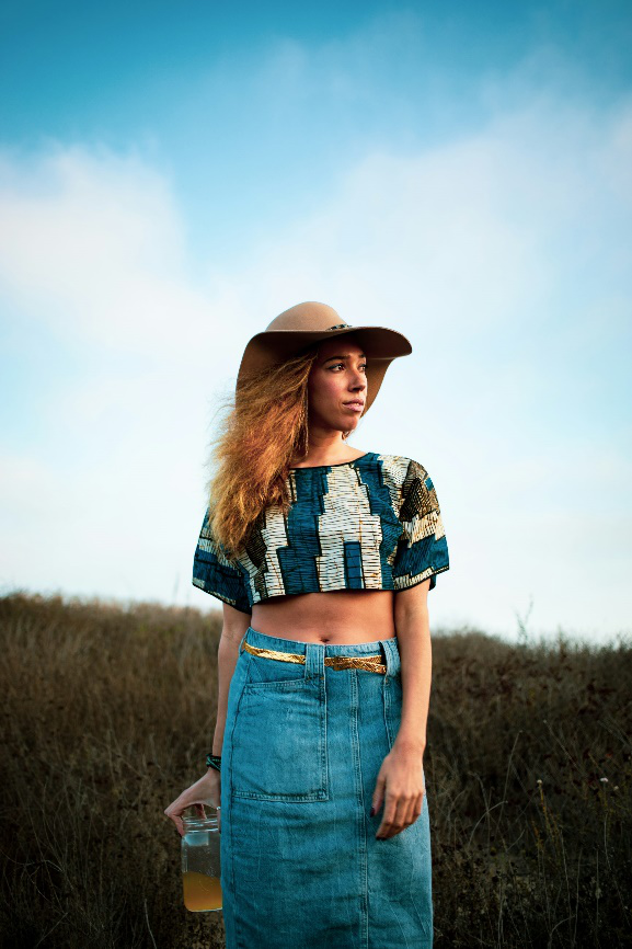 Woman wearing a denim skirt and patterned crop top with a wide-brimmed hat in a field at sunset.