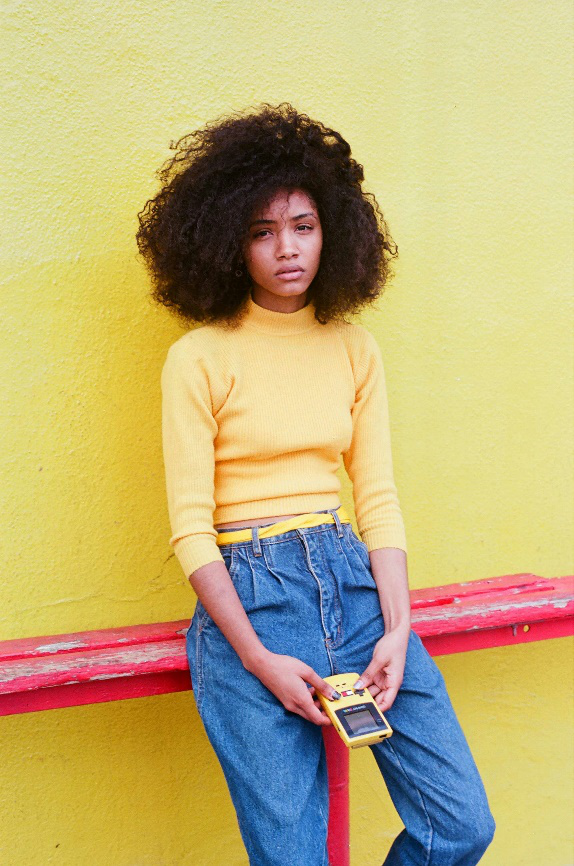 Young woman in a mustard yellow sweater and high-waisted jeans leaning against a bright yellow wall.