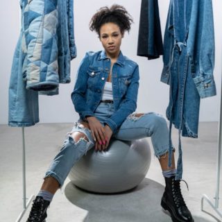 Woman wearing a denim jacket and pants, posing confidently near a clothing rack.