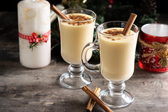Two glasses of Ponche de Creme with cinnamon sticks beside a holiday candle