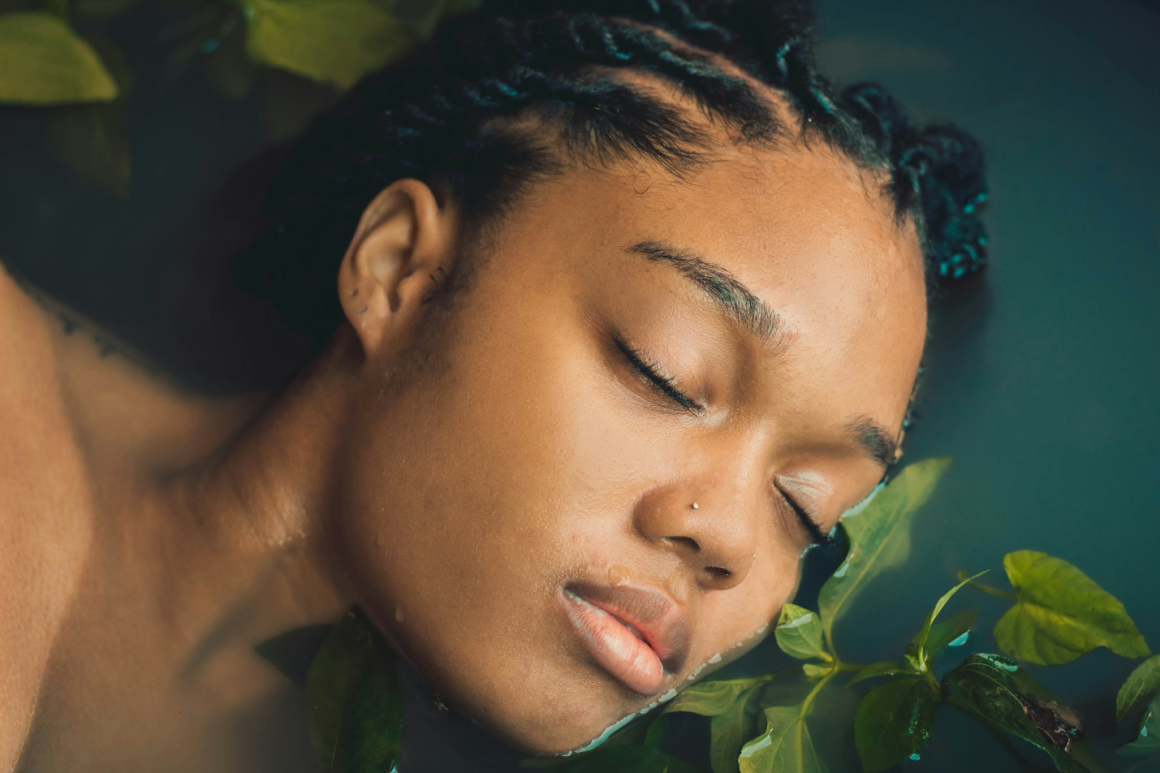 Close-up of a woman resting with her eyes closed and face dipping in water with a plant touching her face.